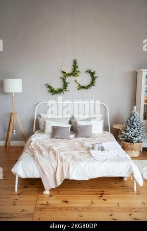 White wrought iron bed with textured milky throws and pillows on a wood floor next to a Christmas tree. Natural green wreaths with a garland hang on a Stock Photo