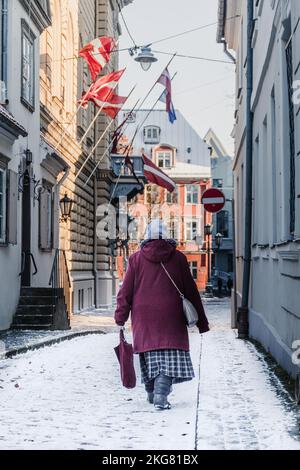 Riga, Latvia - November 20 2022: Elderly woman wearing winter warm clothes walking in a narrow street covered by the snow in Riga Old Town near Parlia Stock Photo