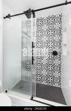 A shower with a black and white patterned tile on the wall, black circular tiles on the floor, and a glass sliding door. Stock Photo