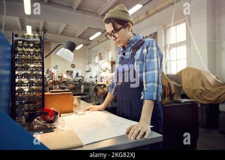 Serious concentrated young woman in workwear standing at table and watching assembly plan of watch movement Stock Photo