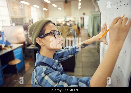 Side view of focused young woman in glasses standing at factory shop and drafting technical sketch leaned to wall Stock Photo