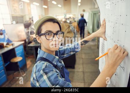 Portrait of serious skilled factory worker in eyeglasses standing at wall in factory shop and analyzing engineering blueprint Stock Photo