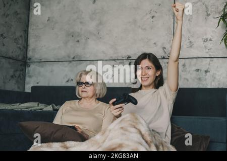 Happy smiling adult daughter and senior mother playing video game using game pad at home. Concept of entertainment, family bonding and leisure Stock Photo