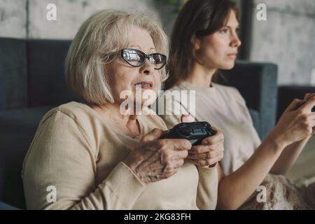 Daughter and mother enjoy playing video game together sitting on sofa in cozy home, smiling and laughing together. Happy family concept. Stock Photo