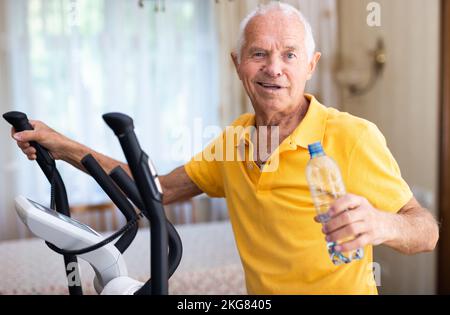 Senior man with bottle of water using elliptical trainer at home Stock Photo