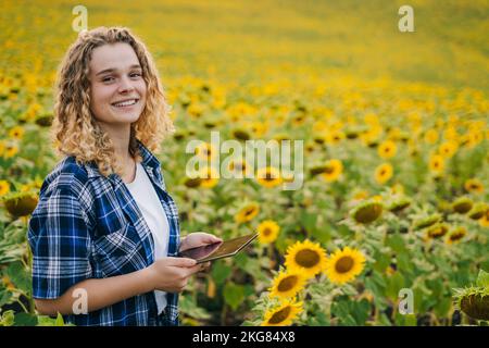 Farmer woman with curly hair looking at camera holding a phone among yellow sunflower fields. Sunflower business concept. Smart farm. Copy space Stock Photo