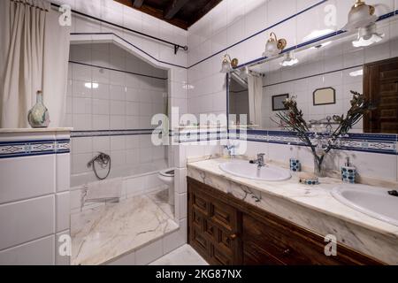 Vintage stately-style bathroom with marble countertops, twin sinks, a large mirror with shades, and a bathtub between white tiles Stock Photo