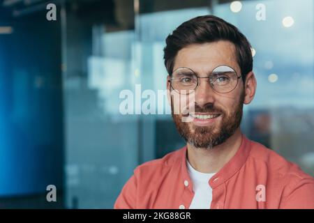 Close-up photo of happy and successful smiling programmer in red casual shirt, man in glasses looking at camera and smiling, developer engineer working inside office. Stock Photo