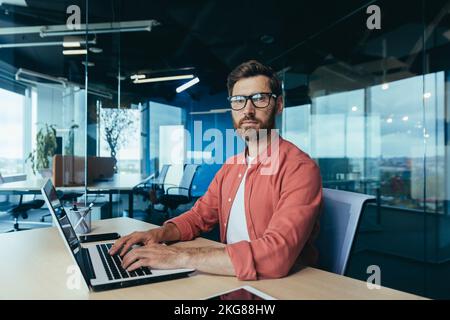 Portrait of a successful programmer inside the office, a man with a beard, glasses and a red shirt looks at the camera, a developer engineer uses a laptop to write code in a programming framework. Stock Photo