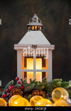 A candle burns in a white lamp on a black background. In the foreground lies a spruce branch and a garland. Stock Photo