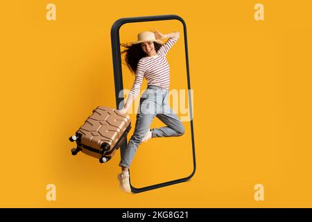 Smiling excited young arab lady tourist in hat with suitcase jumping near huge smartphone, ready for travel Stock Photo