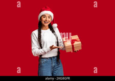 Holiday Bonus. Beautiful Arab Woman In Santa Hat Holding Wrapped Gift Box Stock Photo