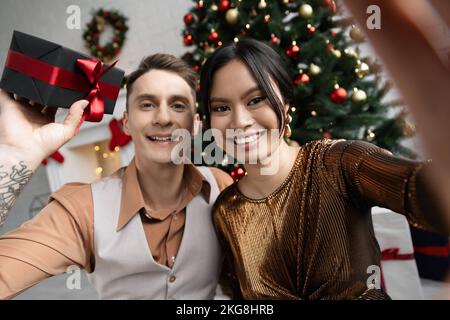 happy tattooed man holding present near asian wife while sitting under Christmas tree Stock Photo