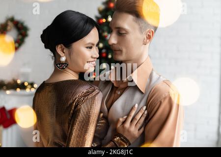 brunette asian woman in festive clothes smiling near young husband during Christmas celebration at home Stock Photo