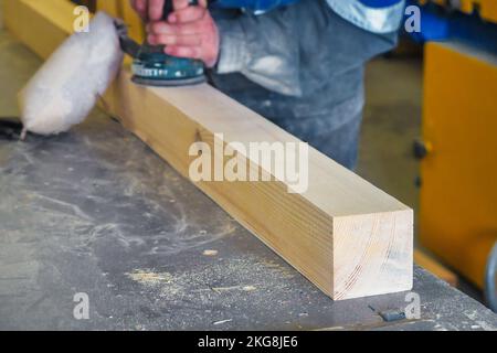 Professional woodworker grinds wooden bar on workbench in carpentry workshop with tool. Middle-aged worker in overalls processes wood with electric grinder. Real workflow.. Stock Photo