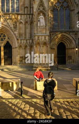 Couple (man woman) by new Elizabeth 2 statue on niche wearing Garter Robes (orb, sceptre) - York Minster front entrance, North Yorkshire, England, UK. Stock Photo