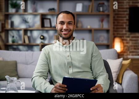 Young African American man sitting at home in a stylish office. Psychologist, coach or businessman looking at the camera and smiling. Beautiful teeth Stock Photo
