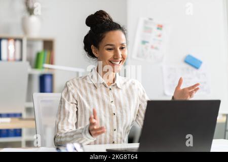 Excited female entrepreneur video calling via laptop, talking to webcamera and smiling, sitting at workplace in office Stock Photo