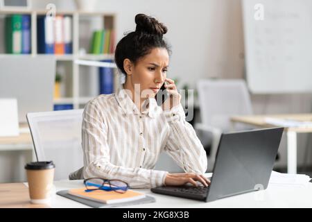 Serious businesswoman talking on smartphone and typing on laptop keyboard, sitting at workplace in office interior Stock Photo