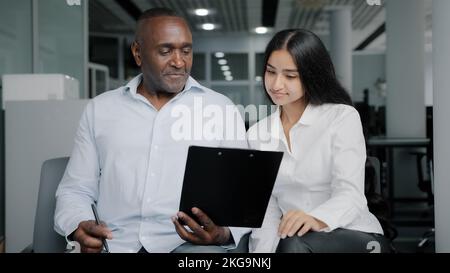 Diverse business partners sit in office discuss paper instruction african businessman show documentation in folder tablet to arabian female colleague Stock Photo