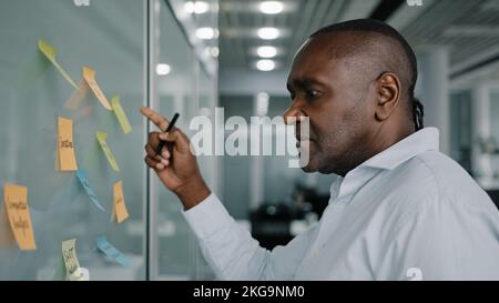 African businessman executive content manager develop strategy plan manage corporate work tasks read sticky notes glued to board write ideas project Stock Photo