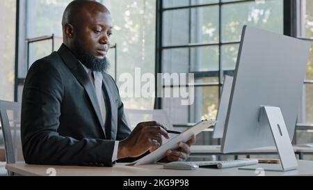 African writer writing notes on paper tablet sit at office desk adult entrepreneur american businessman create new ideas corporate tasks business Stock Photo