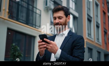 Attractive caucasian businessman stand in city texting messages on mobile phone positive smiling 30s freelancer owner read internet digital news good Stock Photo