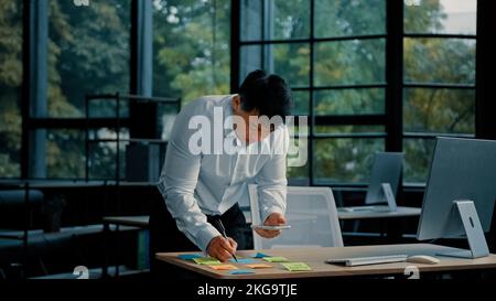 Korean asian employee businessman stand at desk in office write on memo stickers manage project tasks. Professional male designer ethnic adult Stock Photo