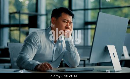 Bored lazy asian worker businessman korean japanese man exhausted weary male sit in office asleep tired from boring online work at computer develop Stock Photo