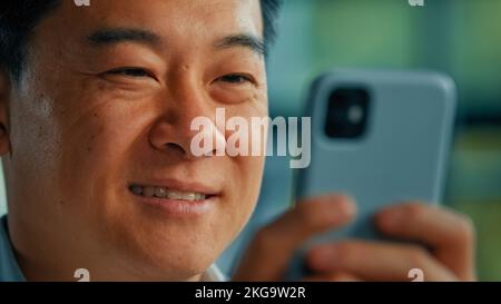 Close up face male user millennial asian adult man businessman positive happy smiling business person send messages on mobile phone at office use Stock Photo