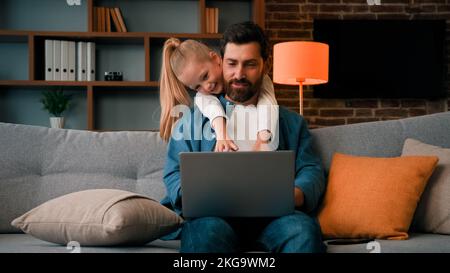 Cute active naughty girl noisy child plays with daddy hugs father distract parental attention disturbs parent from work on laptop at home single Stock Photo