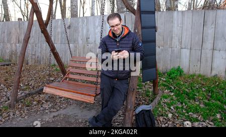Man charging smartphone from portable solar panel outdoor near the swing. Clean energy for using in camping. Work with gadgets outdoor with portable s Stock Photo