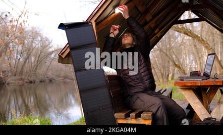 Man work on laptop charginf from portable solar panel utdoor in alclove near the river. Clean energy for using in camping. Work with gadgets outdoor w Stock Photo
