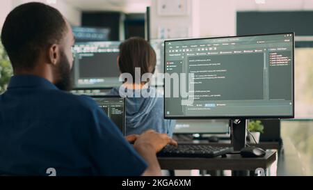 African american system engineer working on software development with source code, looking at computer. IT app developer writing ai algorithm in office, online cloud computing. Handheld shot. Stock Photo