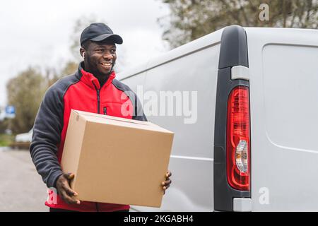 Package delivery concept. Outdoor medium closeup shot of middle-aged African-American handsome deliveryman in red jacket holding a big package and standing in front of white delivery truck. High quality photo Stock Photo