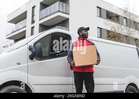 Medium long outdoor shot of tall focused Black deliveryman carrying a big parcel packed in cardboard box standing in front of big delivery truck. Blurred building block in the background. High quality photo Stock Photo