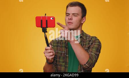 Tourist man in shirt traveler blogger taking selfie on mobile phone selfie stick communicating video call online with subscribers. Young guy boy 20s isolated alone on yellow studio wall background Stock Photo
