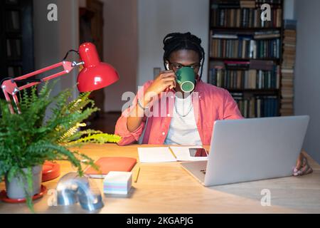Young focused African American man freelancer drinking coffee from mug sits at work desk with laptop Stock Photo