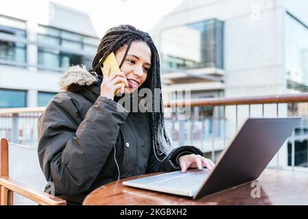 Satisfied Hispanic lady with Afro braids in outerwear speaking on cellphone while sitting at table with laptop in street cafe Stock Photo