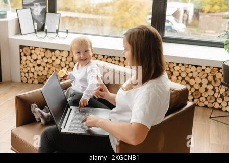 Work at home hybrid job. mother works remotely via laptop next to her son who interferes with her work. Stock Photo