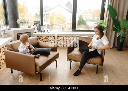 Mother and son work together remotely. The son sits next to her in the chair. A woman works on her laptop at home Stock Photo