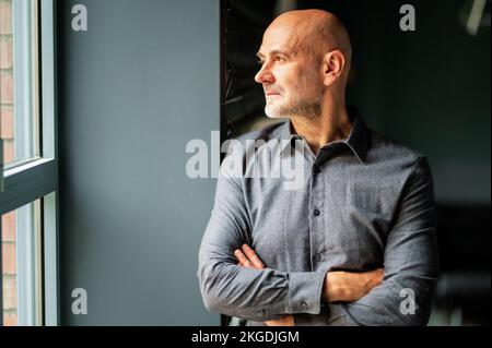 Portrait of middle aged man wearing casual clothes and standing with arms crossed at the window. Confident male looking thoughtfully while looking out Stock Photo