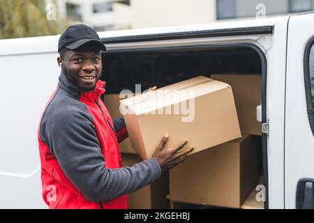 Outdoor medium closeup shot of positive handsome Black man in red jumper with gray sleeves and black baseball hat holding one of many cardboard parcels. Shipping and online shopping concept. High quality photo Stock Photo