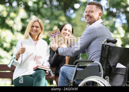 Smiling disabled man in wheelchair studying english with friends and showing thumbup sign Stock Photo