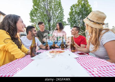 people having fun around a wooden table with take away pizza and bottles of beer, young group of couples singing and playing music to feel carefree an Stock Photo