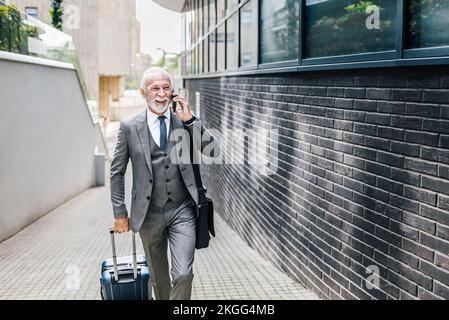Smiling senior businessman with suitcase talking on smart phone. Elderly professional wearing suit is carrying laptop bag. He is walking on footpath b Stock Photo