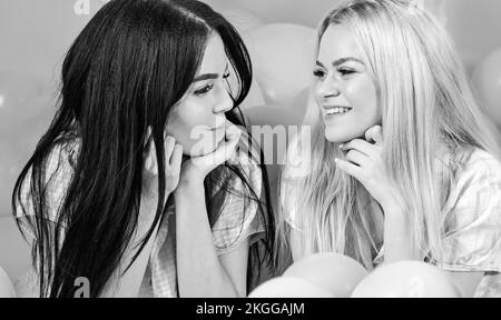 Girls lay on belly near balloons, pink background. Gossip concept. Sisters or best friends in pajamas at girlish pajamas party. Blonde and brunette on Stock Photo
