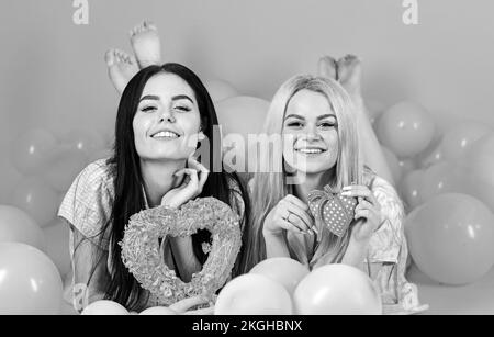 Girls lay near balloons, holds heart toys, pink background. Sisters, friends in pajamas at pajamas party. Blonde and brunette on smiling faces Stock Photo