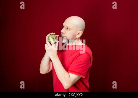 Young handsome man with beard, bald head and red shirt standing holding pinecone fruit against red background. Stock Photo