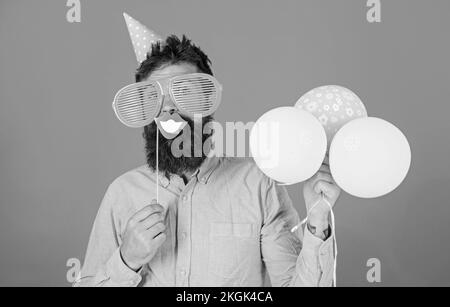 Guy in party hat with air balloons celebrates. Hipster in giant sunglasses celebrating. Man with beard on cheerful face holds smiling mouth on stick Stock Photo